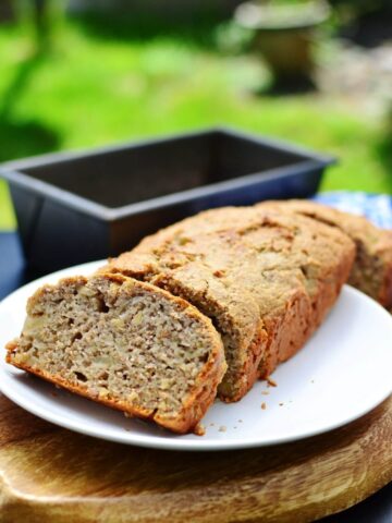 Banana bread with 1 slice on white plate on top of wooden board with bread tin in background.