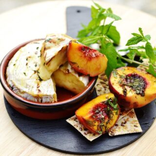 Baked camembert in round brown dish, with halved peaches, crackers and herbs on top of small slate platter.