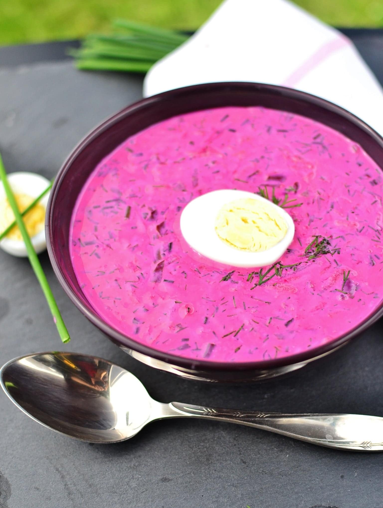 Beet soup with dill and egg half in purple bowl with spoon in front, eggs, chives and white cloth in background.