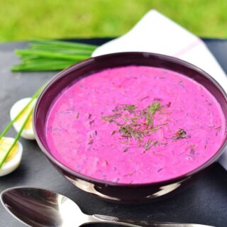 Cold beet soup in purple bowl with spoon in front, boiled eggs, chives and white cloth in background.