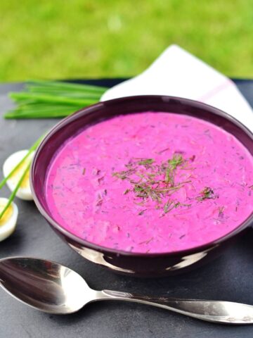 Cold beet soup in purple bowl with spoon in front, boiled eggs, chives and white cloth in background.