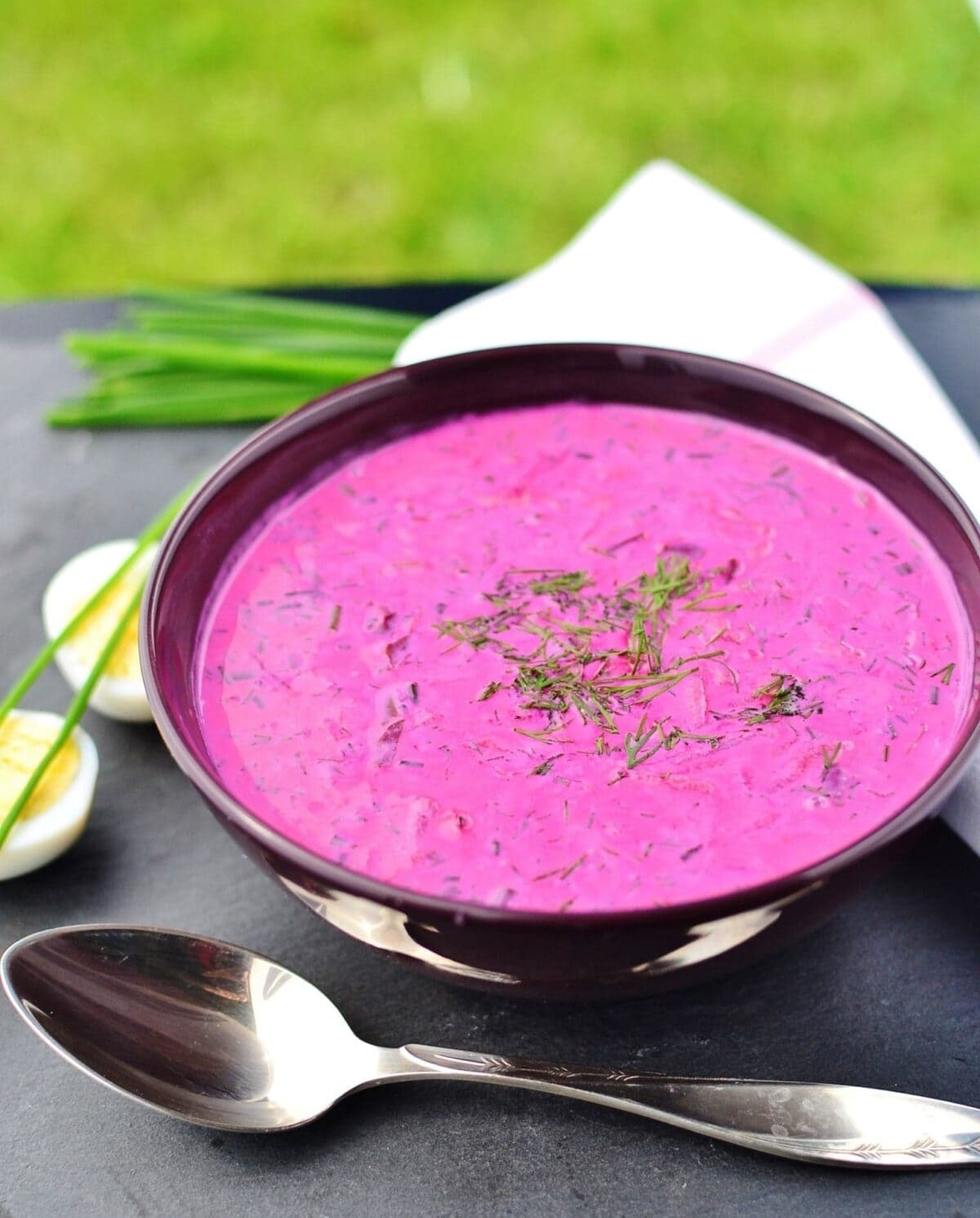 Beet soup garnished with dill in purple bowl with spoon in front, halved eggs, chives and white cloth in background.