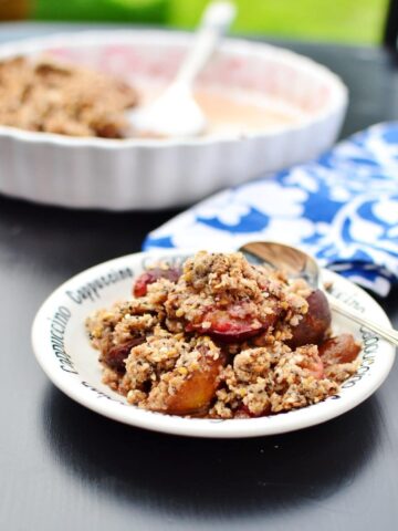 Side view of plum crumble on small white plate with spoon, blue-and-white cloth and white pie dish in background.