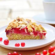 Side view of almond redcurrant cake slice on white plate with redcurrants on wooden surface.