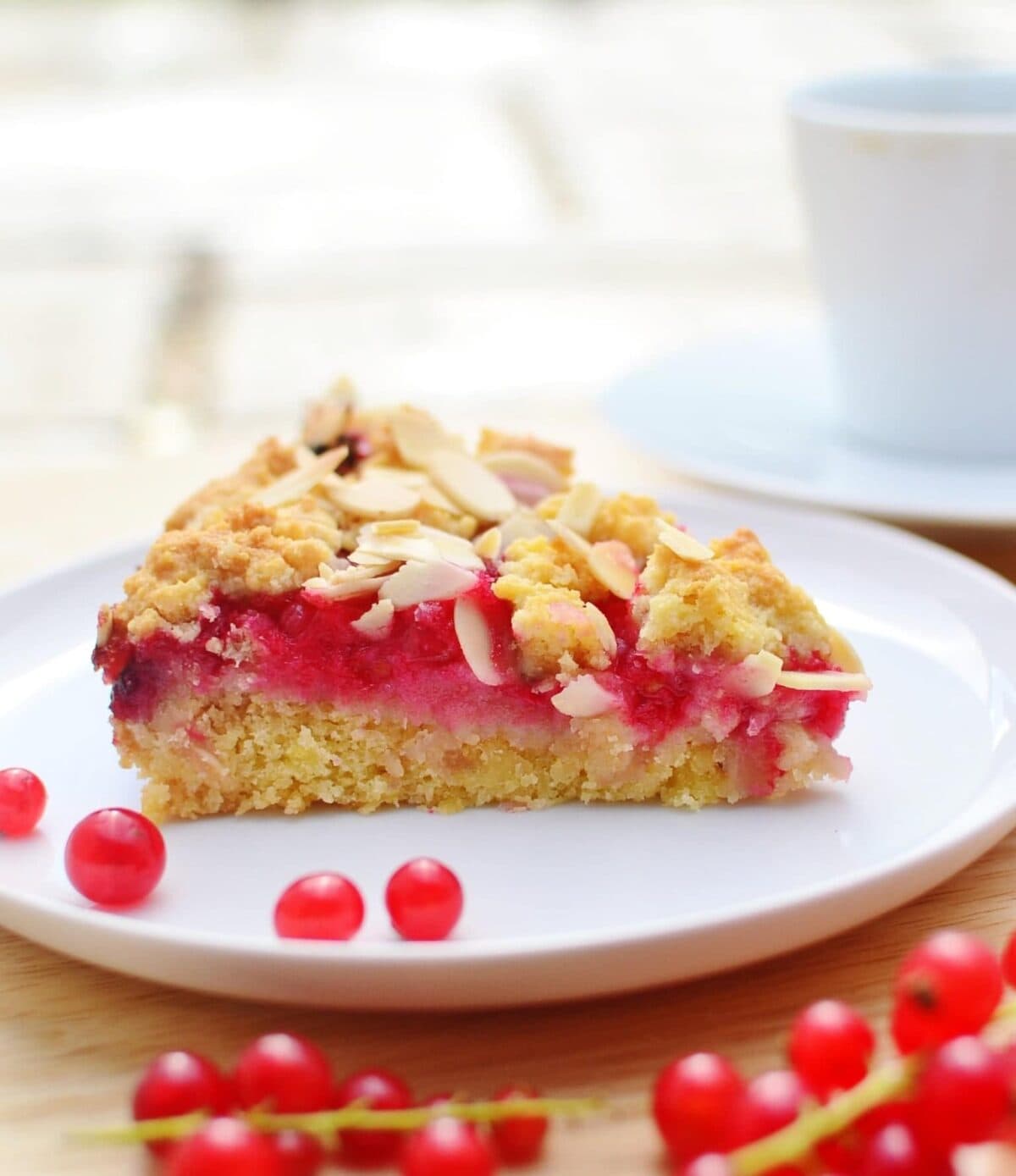 Side view of almond redcurrant cake slice on white plate with redcurrants on wooden surface.