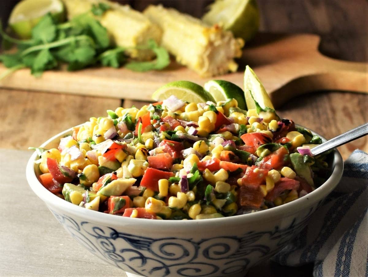 Side view of corn salsa with vegetables in bowl, with corn on cob and herbs in background.