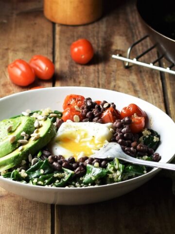 Side view of quinoa breakfast bowl with spinach, sliced avocado, egg, baby tomatoes and fork, with tomatoes, skillet and green cloth with knife in background.