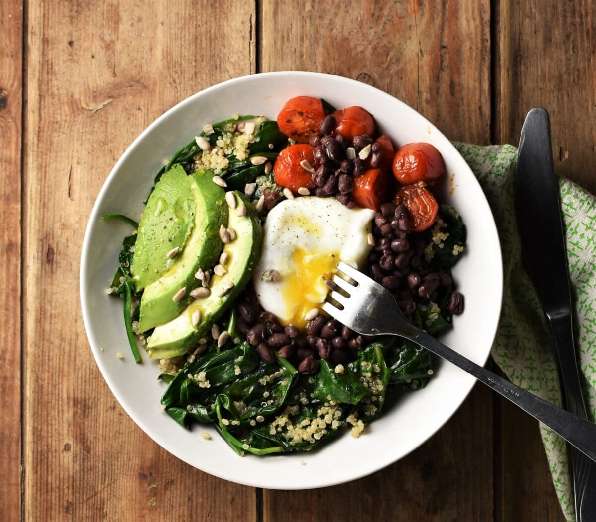 Spinach with quinoa mixture topped with sliced avocado, poached egg, beans and small tomatoes in white bowl with fork and green cloth with knife to the right.