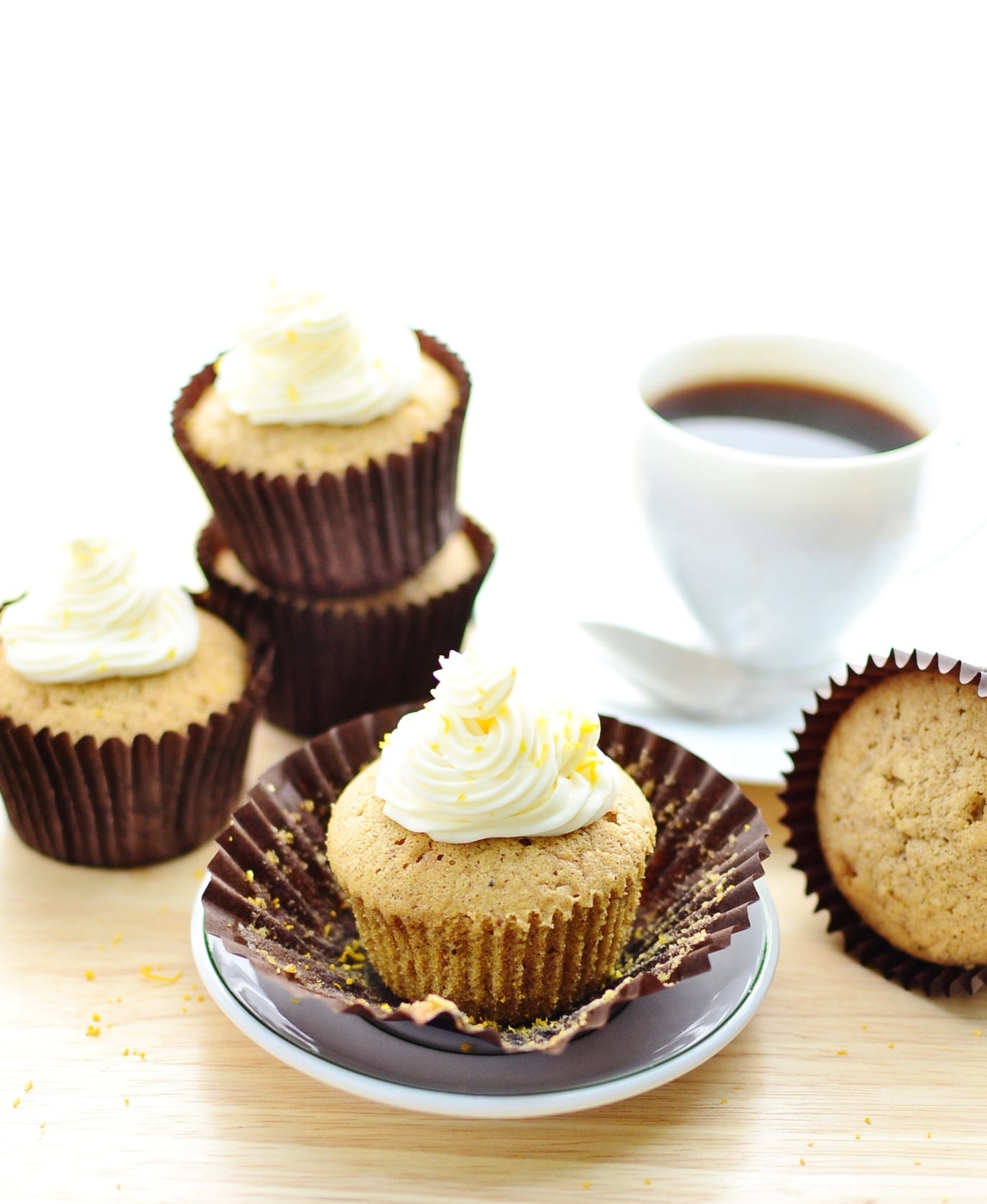 Cupcakes with white frosting in brown paper cases with saucer and white cup with coffee in background.