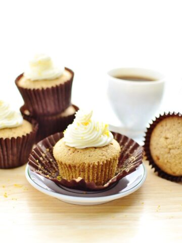 Cupcakes with white frosting in brown paper cases with saucer and white cup with coffee in background.