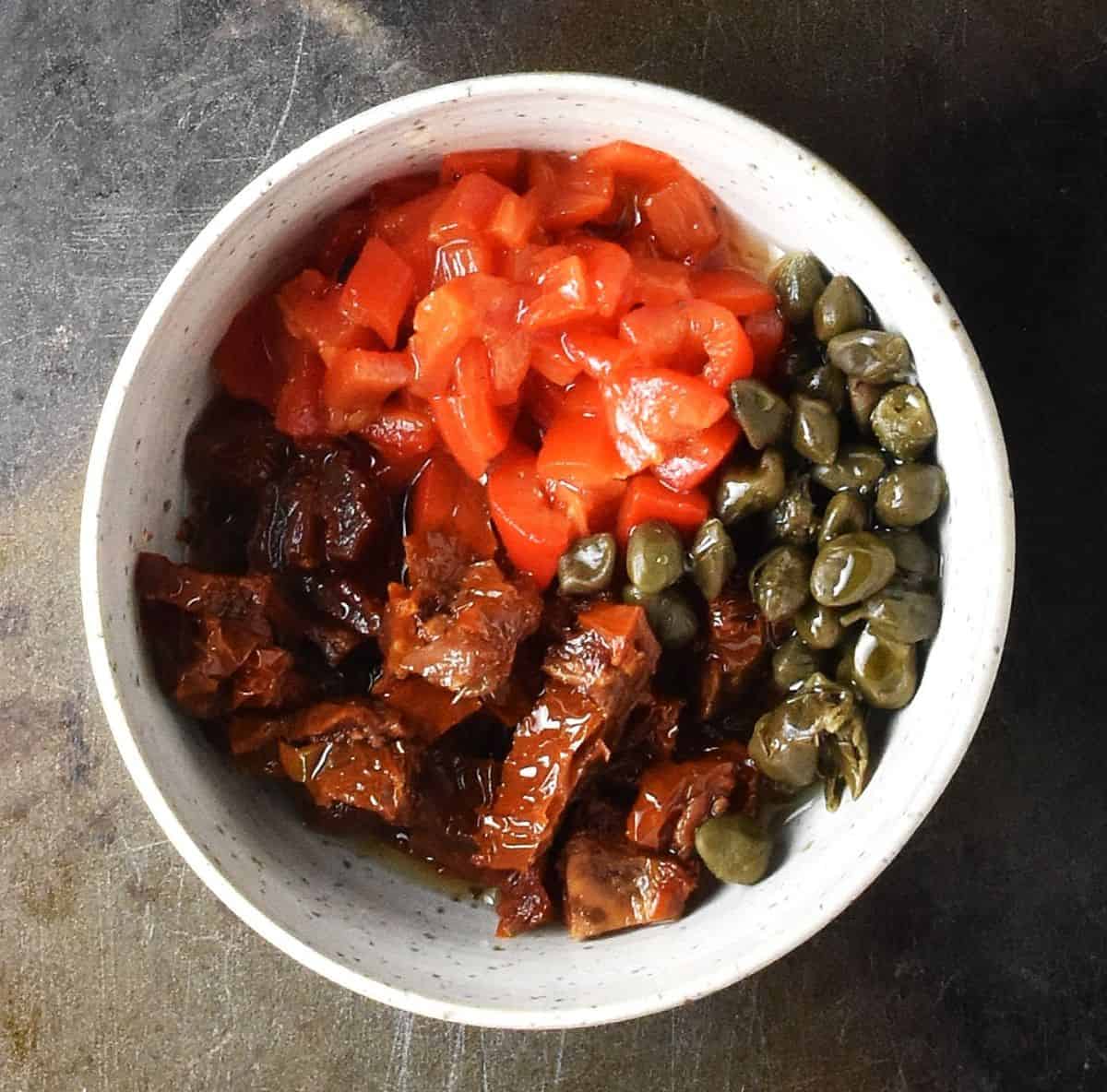 Top down view of chopped sun-dried tomatoes, red pepper and capers in white bowl.