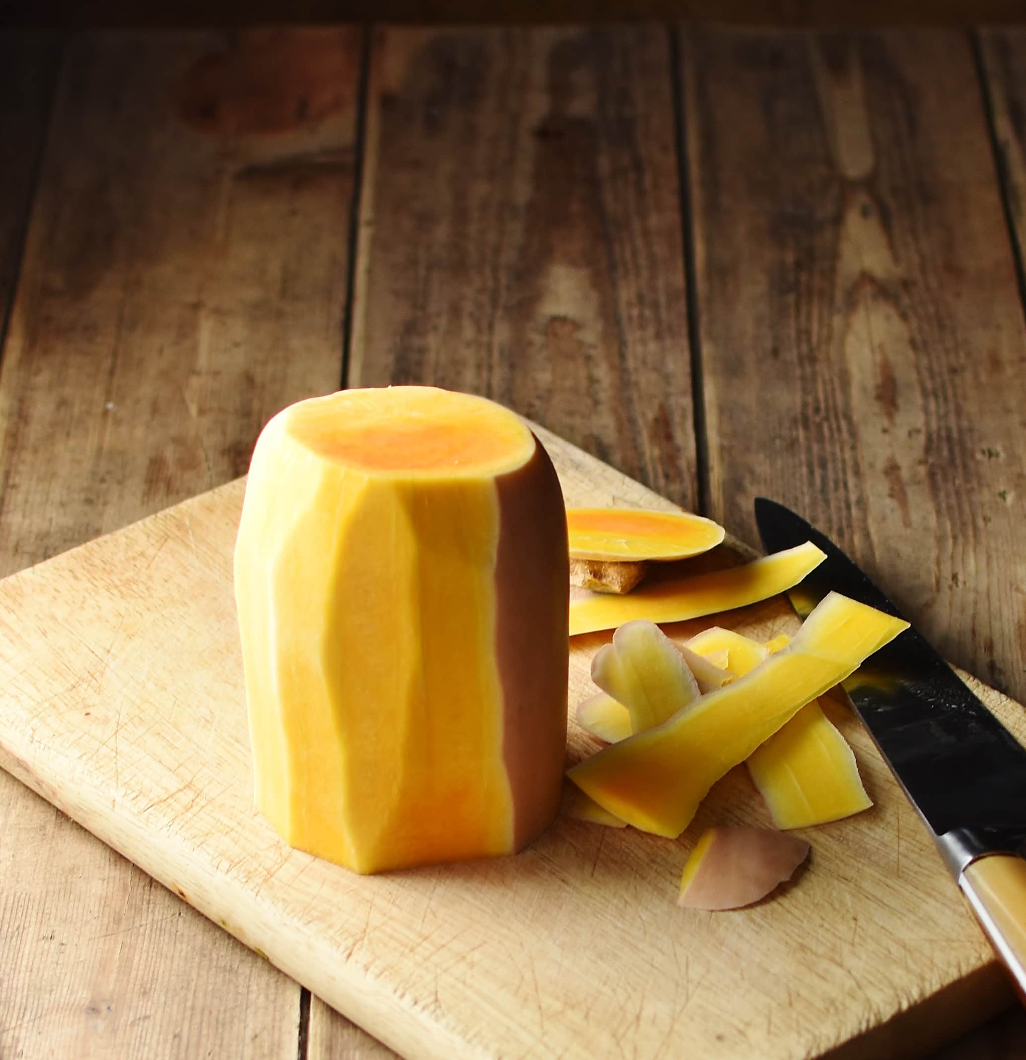 Halved butternut squash partly peeled on top of cutting board with knife.