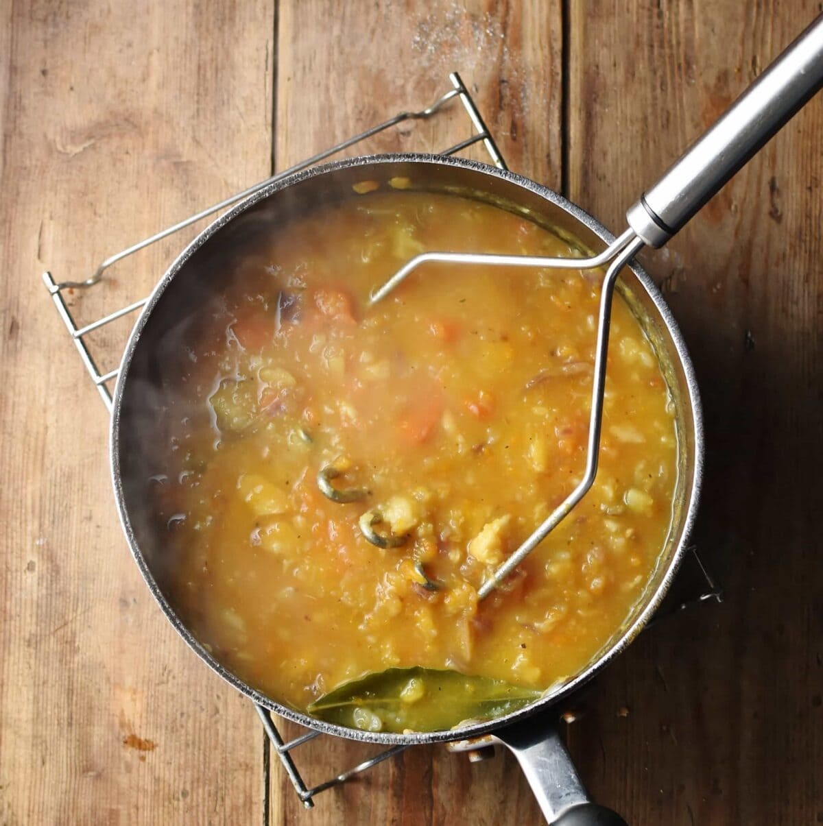 Chunky thick soup with potato masher in large pot on cooling rack.