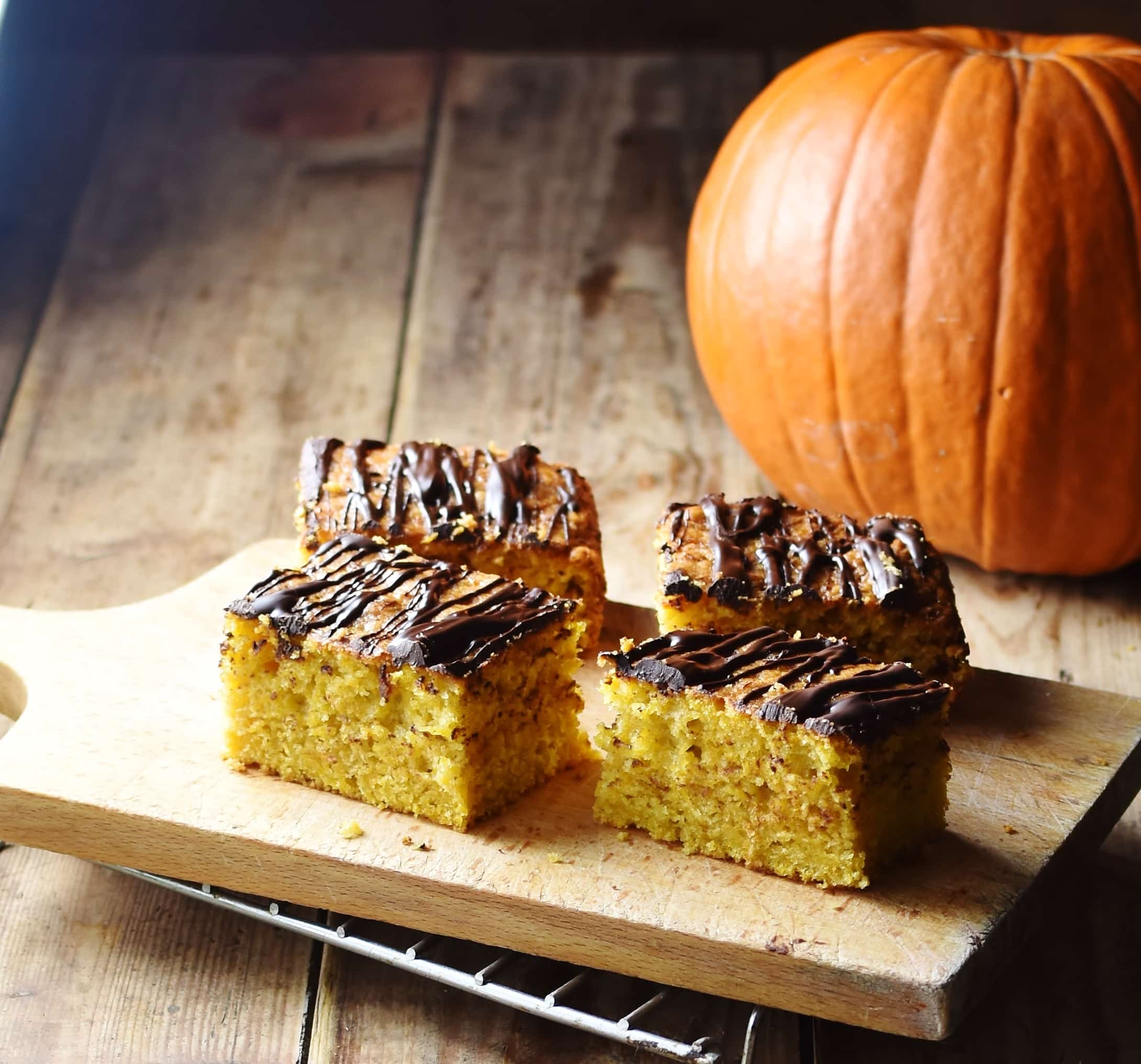 4 slices of pumpkin cake with chocolate drizzle on top of cutting board, with pumpkin in background.