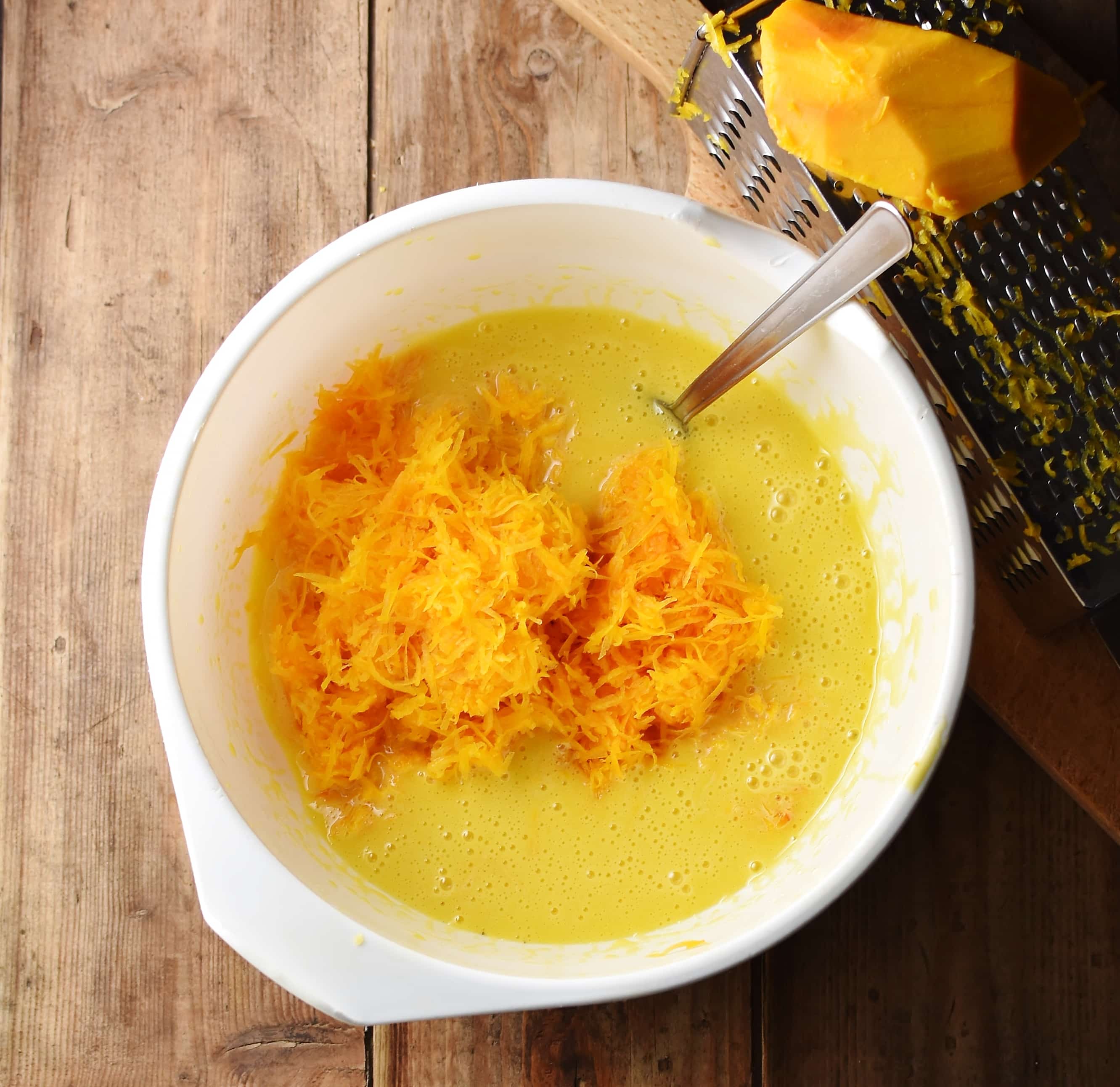 Grated pumpkin and egg mixture in large white bowl with spoon, and grater with pumpkin piece in top right.