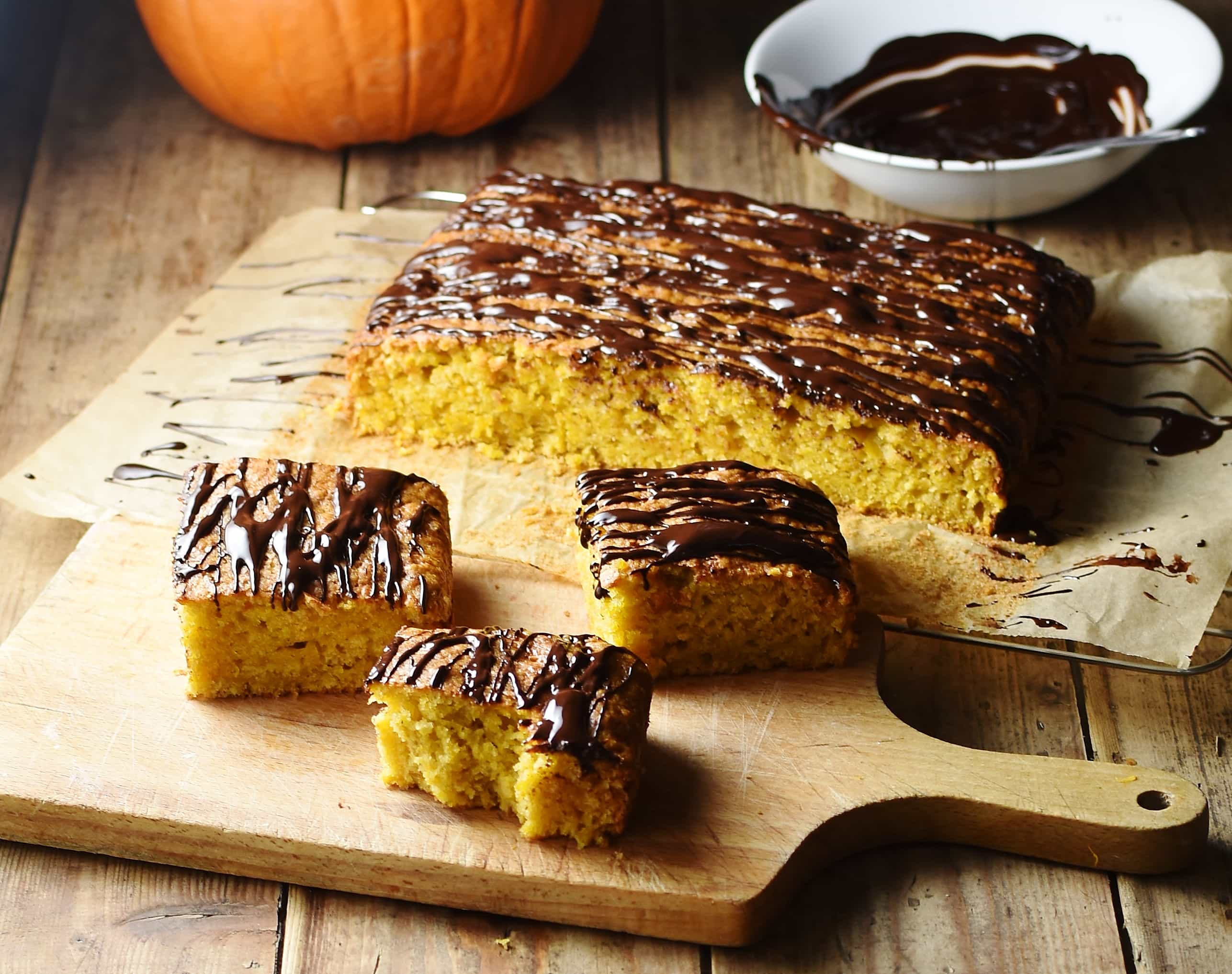 Side view of healthy pumpkin cake slices on top of cutting board, with cake, pumpkin and melted chocolate in white bowl in background.