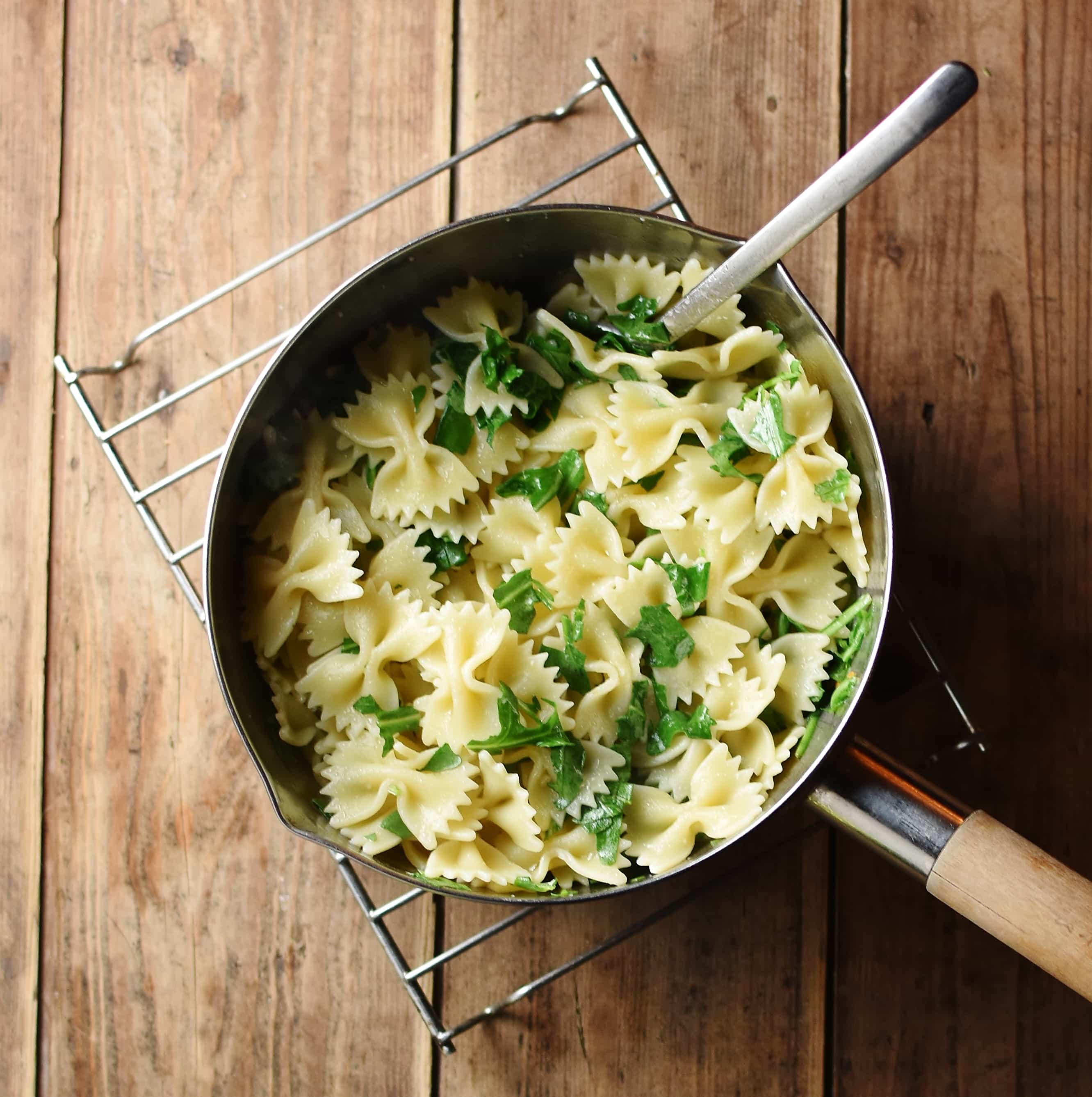 Farfalle pasta with chopped arugula inside saucepan with spoon on cooling rack.