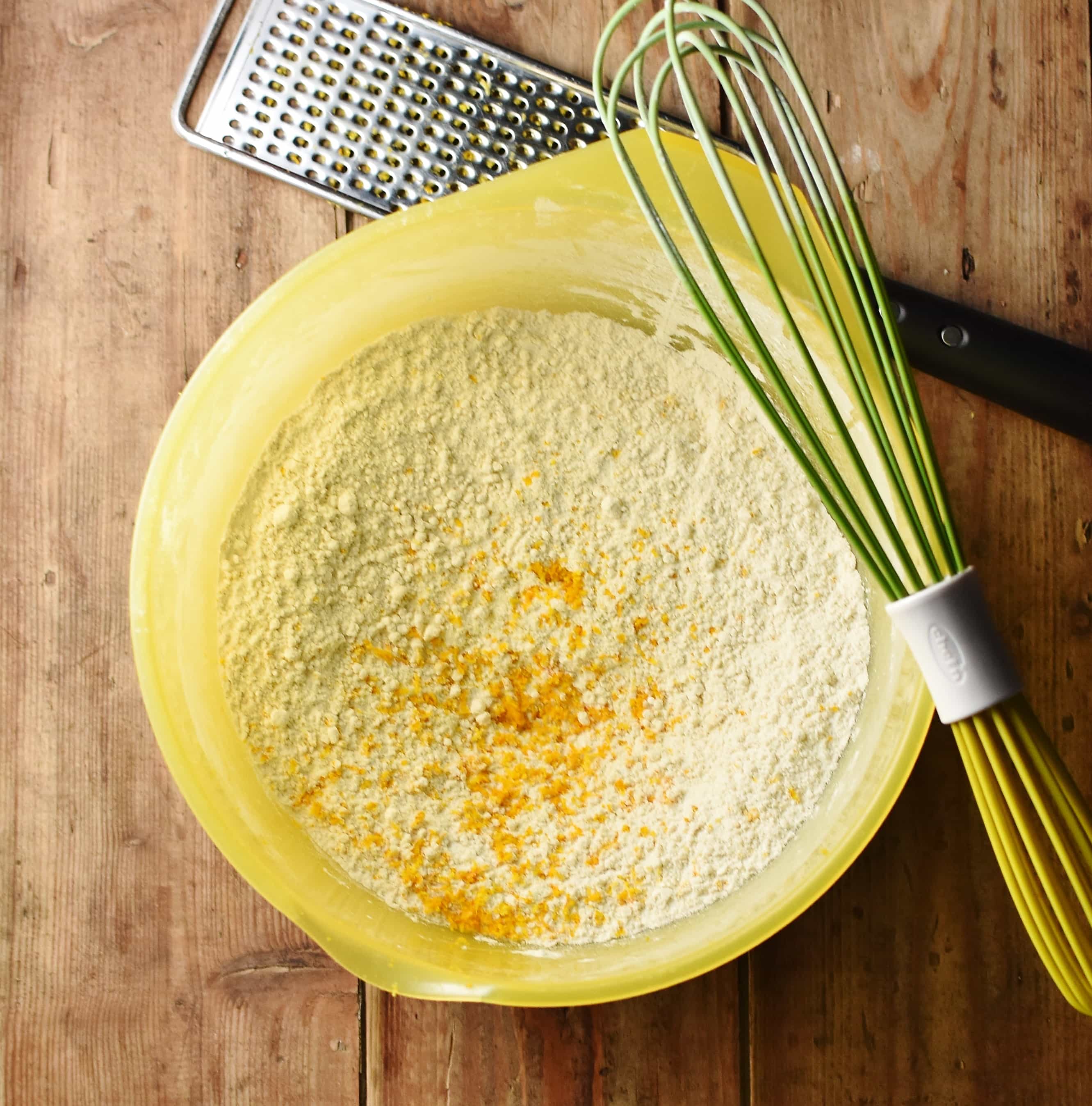 Flour mixture and orange zest in large yellow bowl with green whisk.