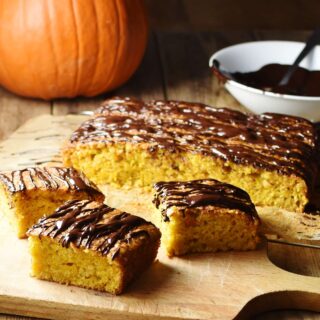 Pumpkin cake slices with chocolate drizzle and more cake in background, with pumpkin and melted chocolate in white bowl in top right.