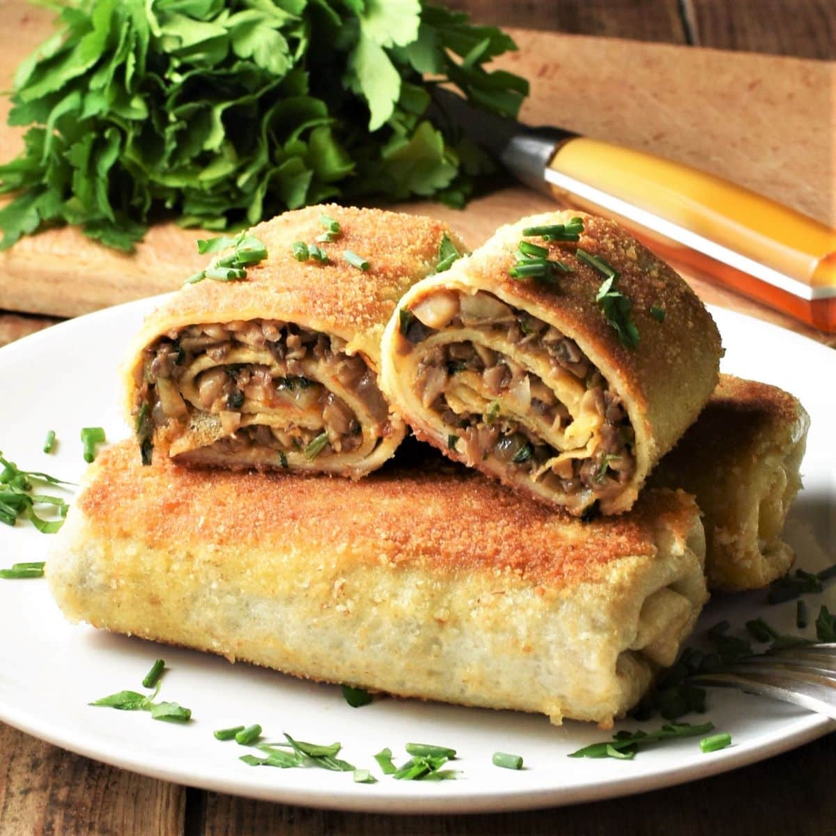 Side view of fried crepes with mushrooms on top of plate with herbs in background.