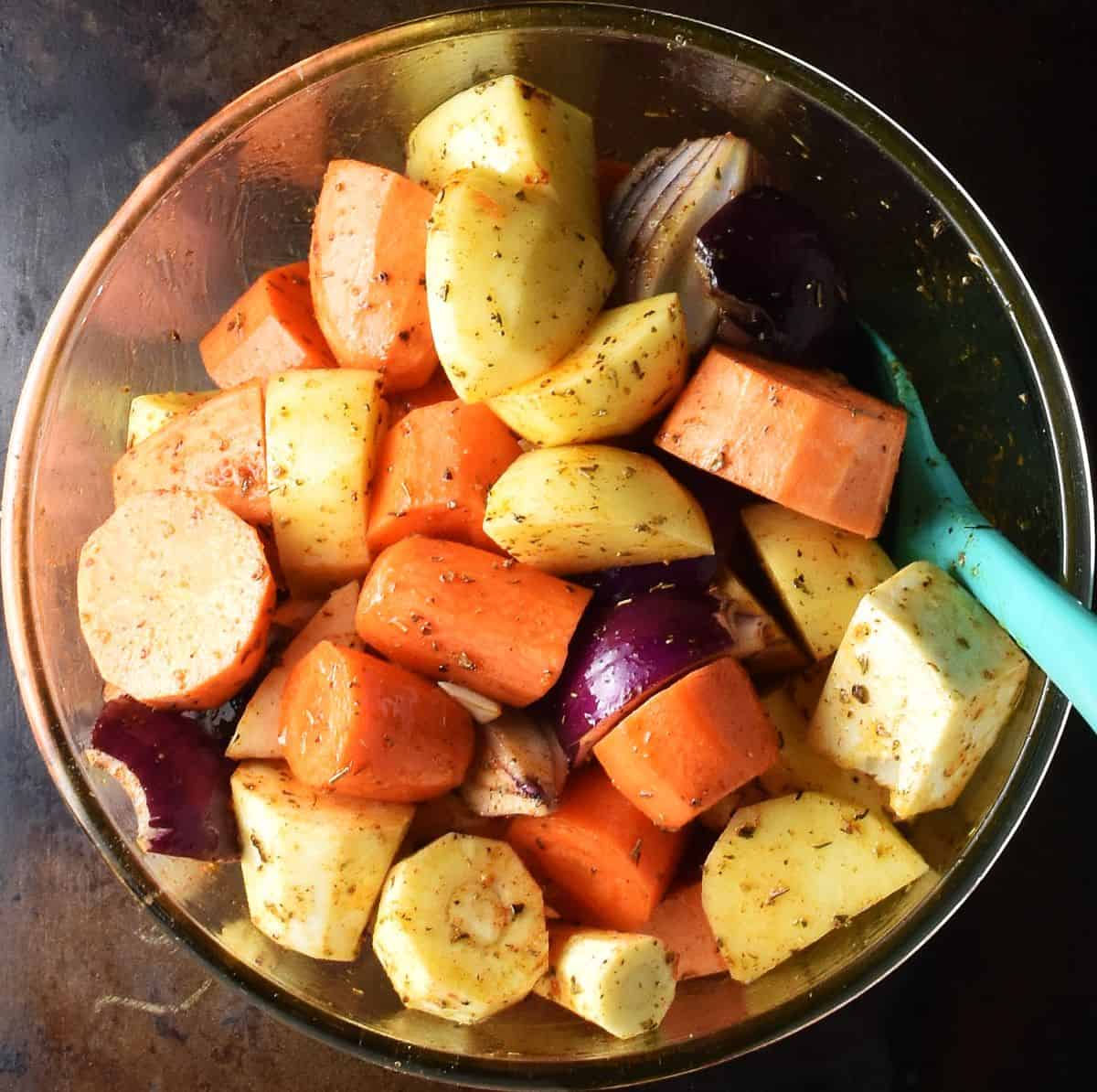 Pieces of root vegetables coated in spices and oil in glass bowl with blue spoon.