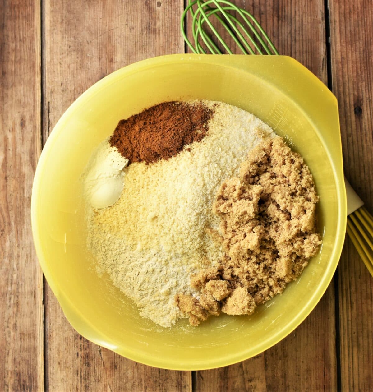 Flour, sugar and spices in large yellow bowl with green whisk to the right.