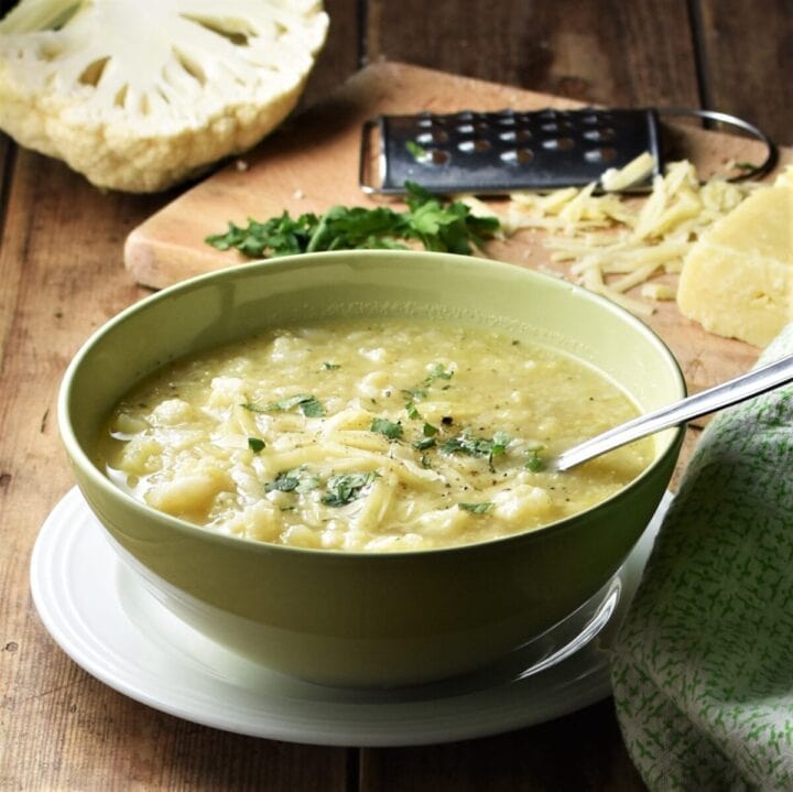 Side view of chunky cauliflower soup in yellow bowl with spoon on top of white plate, with cauliflower, cheese and grater on wooden board, and green cloth in background.