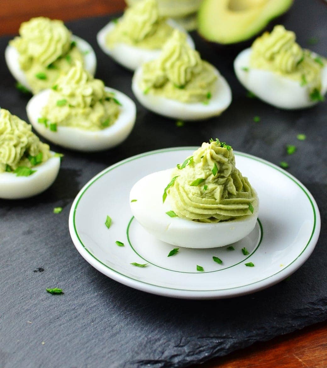 Deviled eggs with avocado and garnish of chives on white plate on top of grey surface.