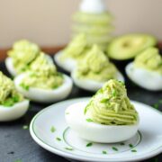Deviled eggs with avocado on small white plate with garnish of chives and deviled eggs in background.