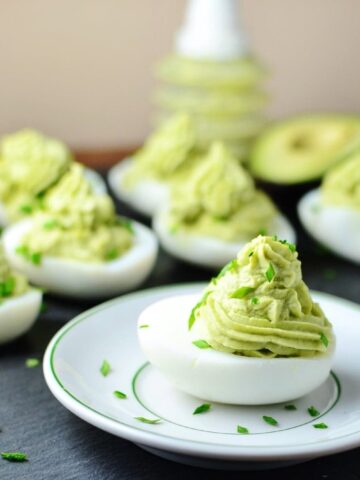 Deviled eggs with avocado on small white plate with garnish of chives and deviled eggs in background.