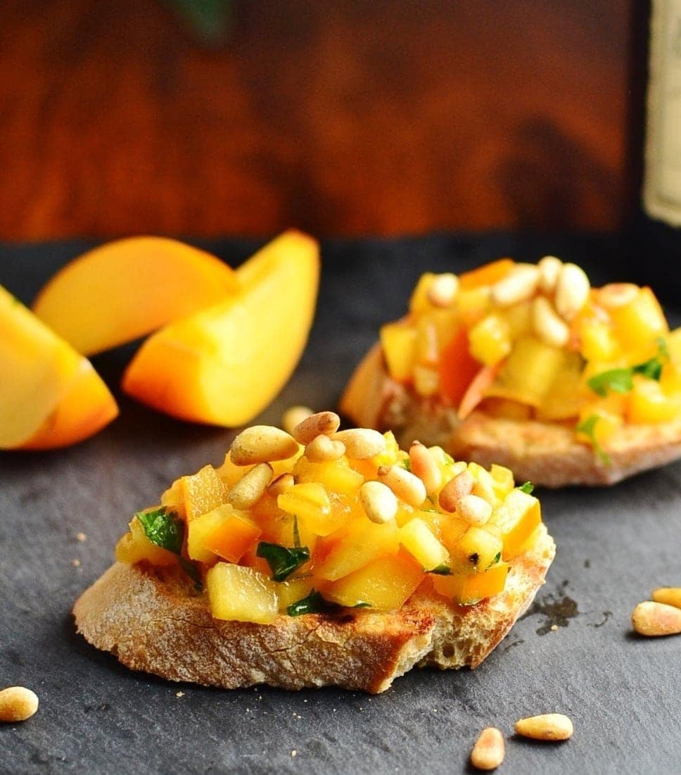 Chopped persimmon mixture on top of 2 baguette slices, with persimmon wedges in background.