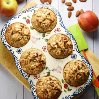Top down view of muffins with almond flakes on top inside 6-hole ceramic muffin pan on top of cutting board, with green spatula, almonds and apples in background.