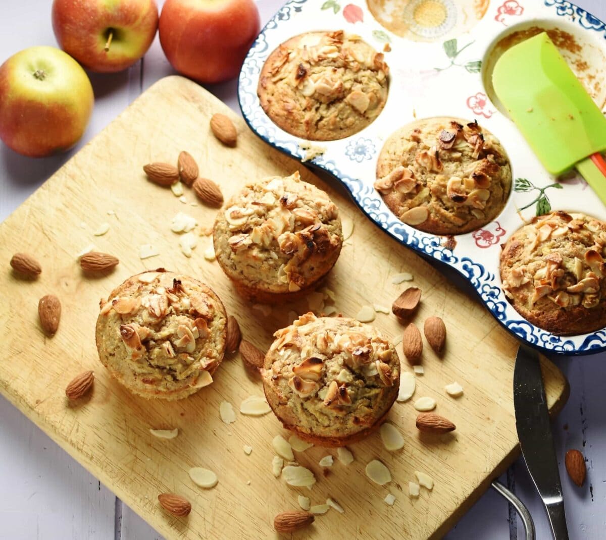 3 muffins with almond flakes on top with almonds on top of wooden board, with 3 more muffins inside ceramic muffin pan and apples in background.