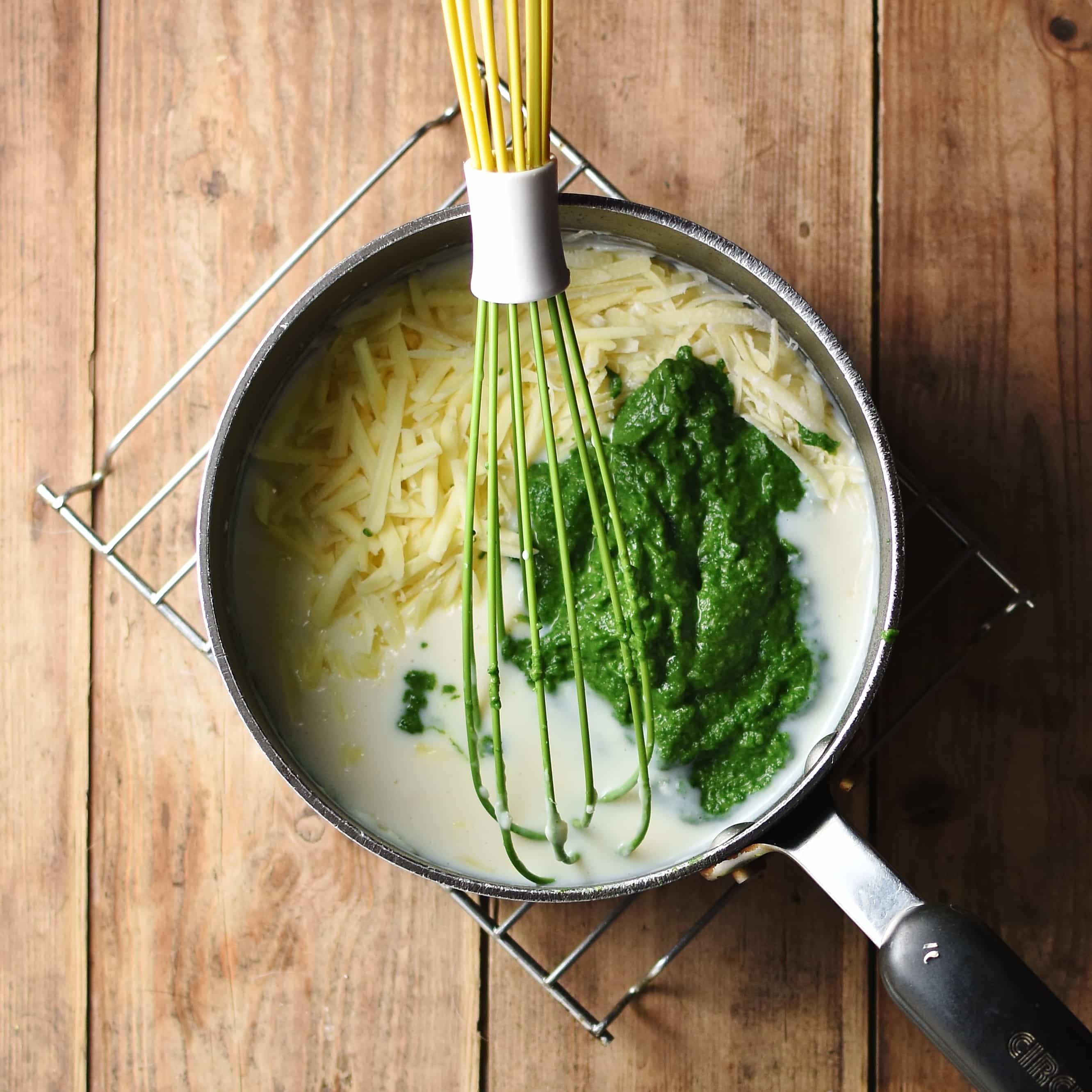 White sauce, pureed spinach and grated cheese in large pot with green whisk.