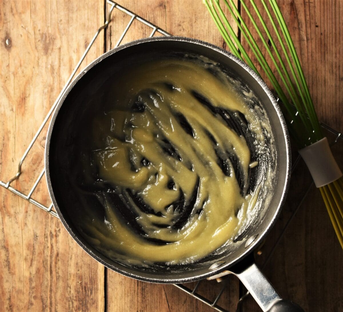 Thick paste (roux) in black saucepan on cooling rack with green whisk in background.