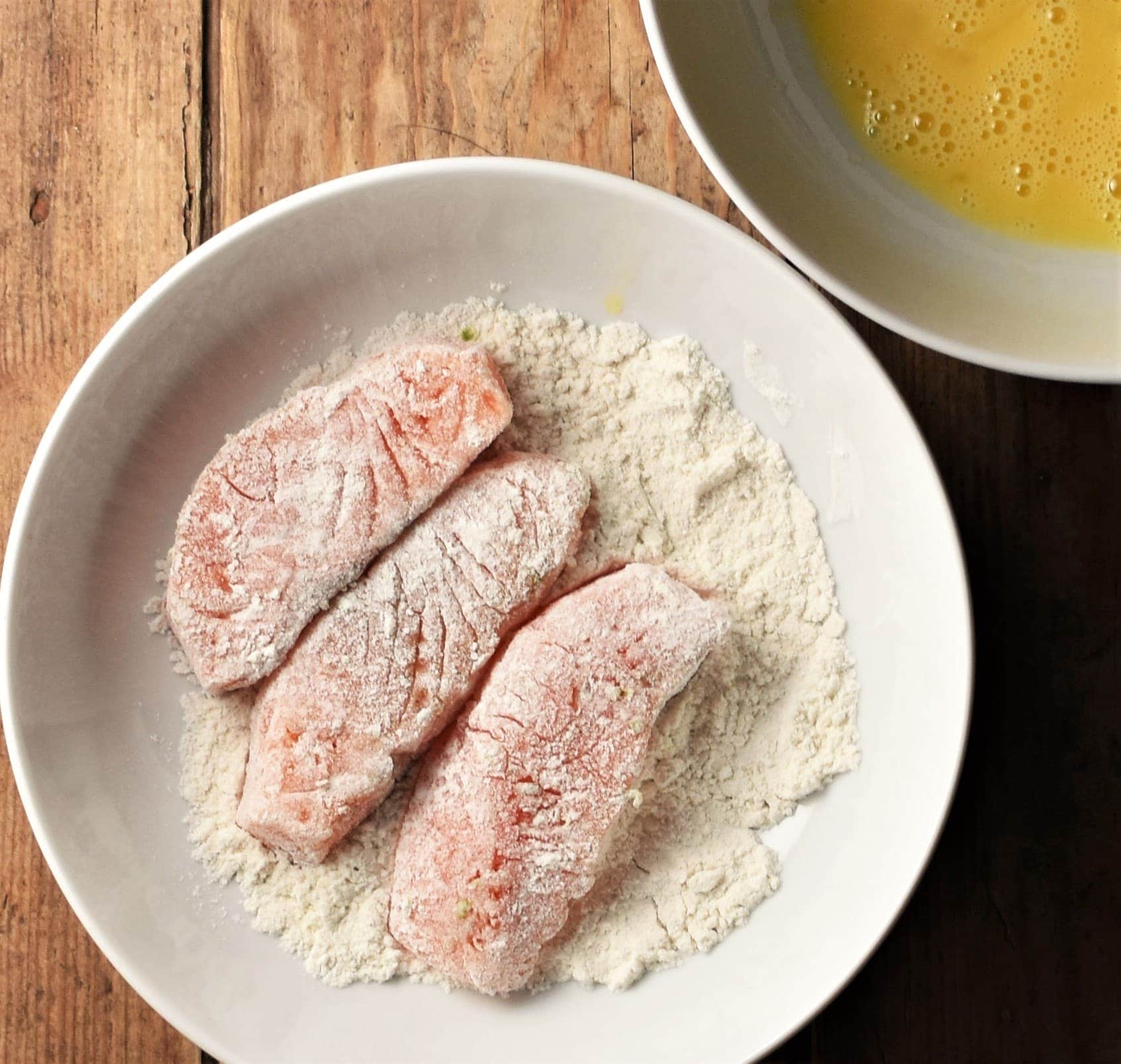 Salmon pieces coated in flour in white bowl with egg wash in background.