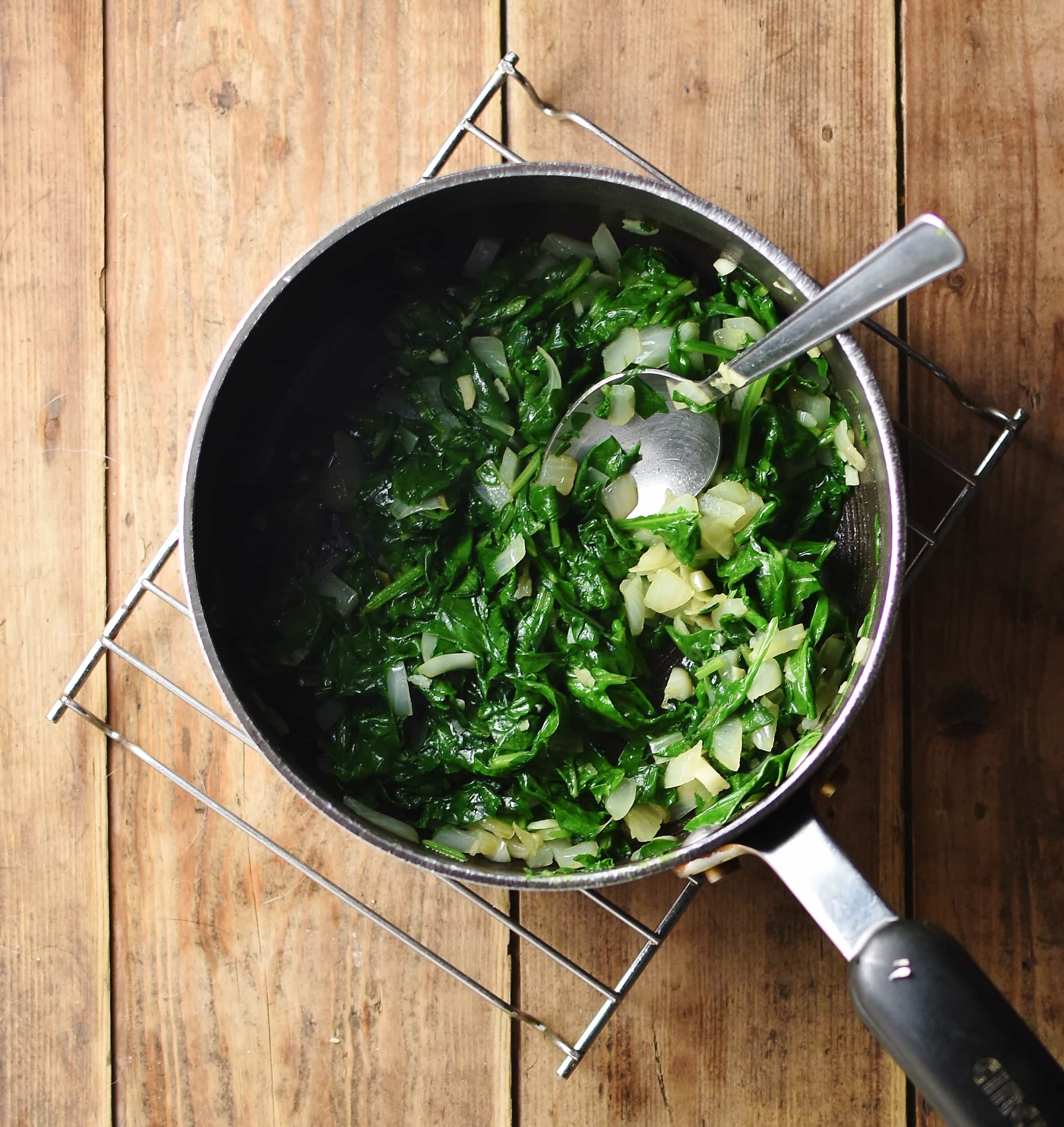 Spinach and onions in large pot with spoon on top of cooling rack.