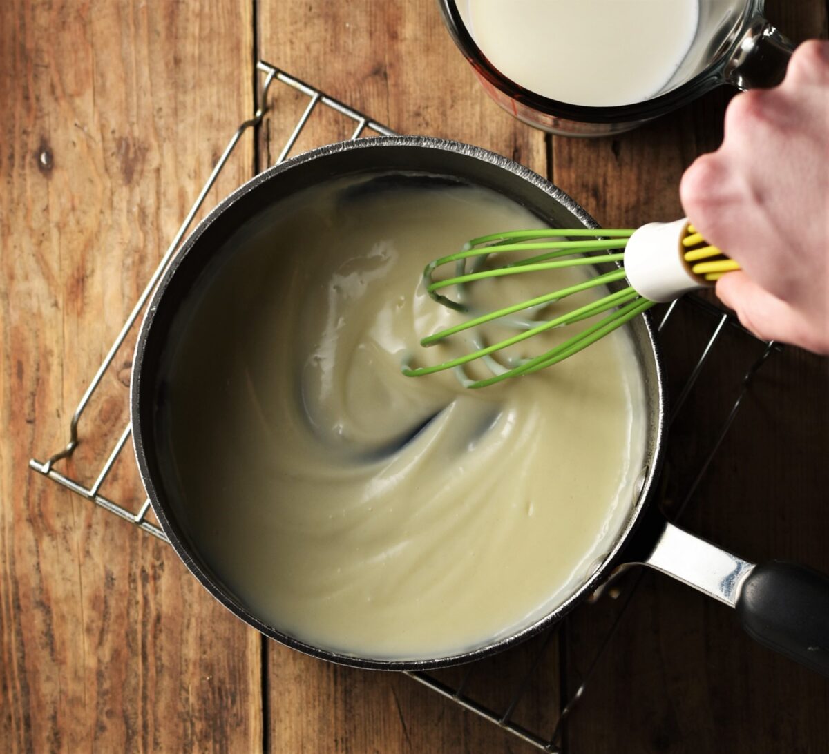 Thick white sauce in black pot with hand holding green whisk on top of rack, with milk in dish at the top.