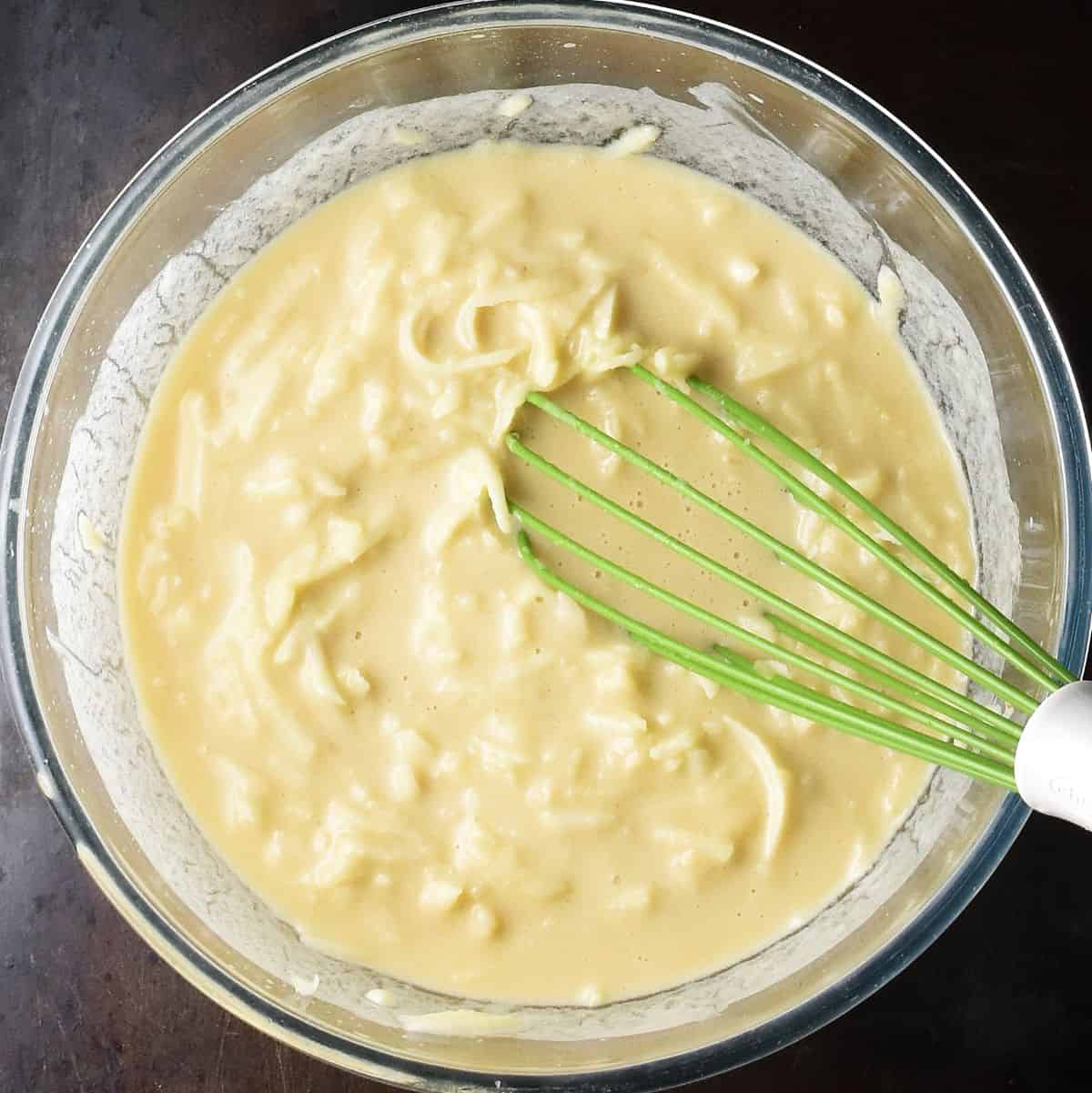 Top down view of grated apple in wet batter in glass bowl with green whisk.
