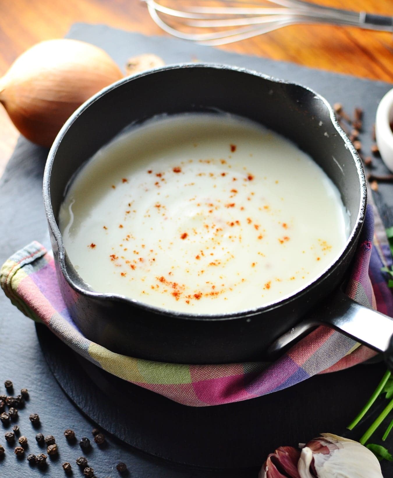 Simple white sauce in black saucepan wrapped in colourful cloth with peppercorns, onion and garlic cloves scattered around.