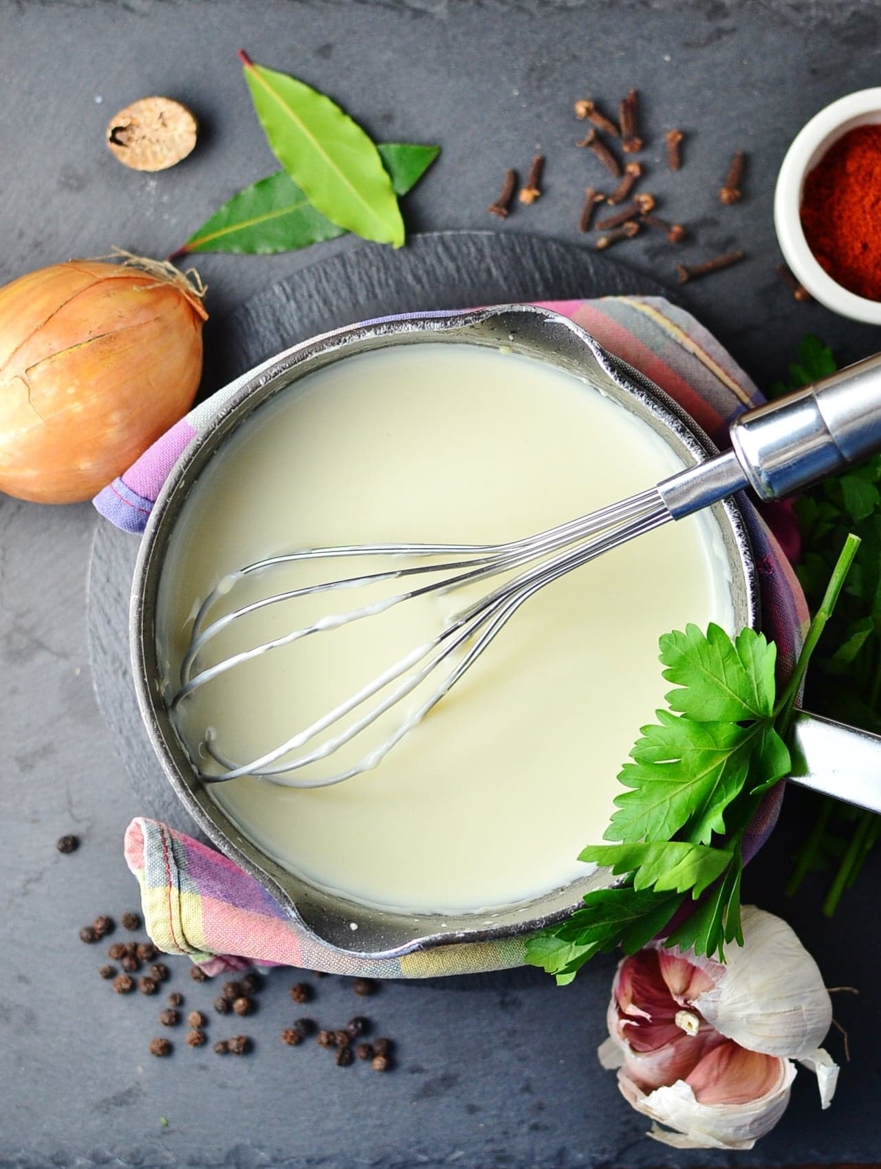 Top down view of bechamel white sauce in saucepan wrapped in cloth, with whisk, onion, garlic and spices around saucepan.