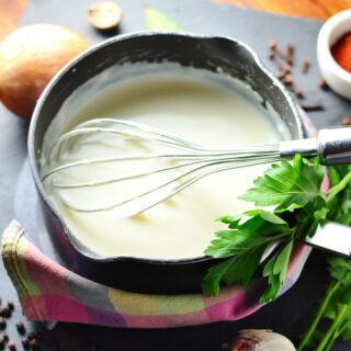 White sauce in black saucepan wrapped in colourful cloth, with whisk, fresh parsley, and peppercorns, onion and garlic cloves scattered around.