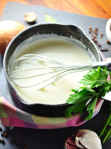 White sauce in black saucepan wrapped in colourful cloth, with whisk, fresh parsley, and peppercorns, onion and garlic cloves scattered around.