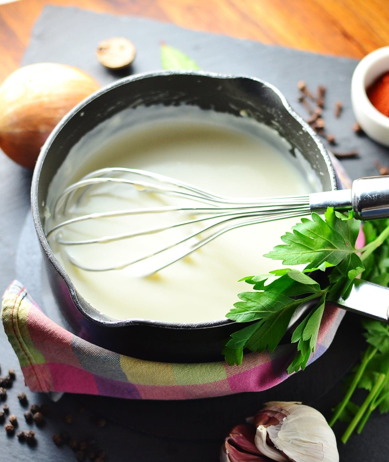 Simple white sauce in black saucepan wrapped in colourful cloth, with whisk, fresh parsley, and peppercorns, onion and garlic cloves scattered around.