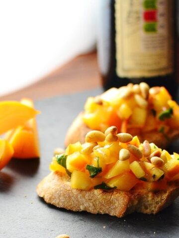 Chopped persimmon and pine nuts on top of 2 baguette slices with bottle in background and persimmon wedges to the left.