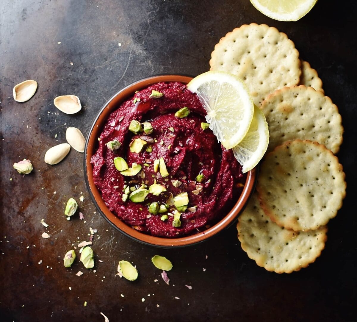 Beet dip with chopped pistachios and lemon wedges inside brown dish, with crackers and pistachios scattered around.