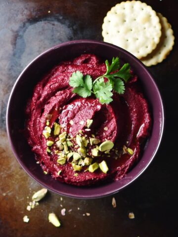 Beetroot dip with pistachios and herbs inside purple bowl, with crackers in background.