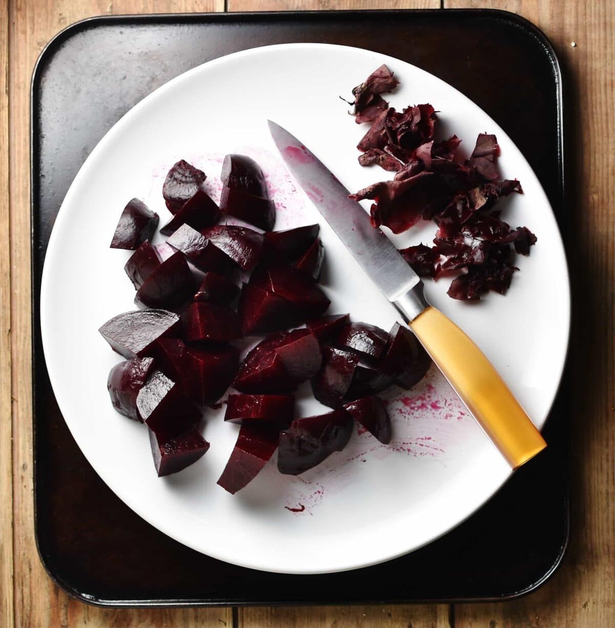 Top down view of chopped beets with peel and small knife on top of white plate.