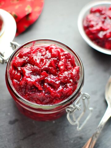 Top down view of cranberry sauce in open jar, with more cranberry sauce in background and spoon in bottom right.