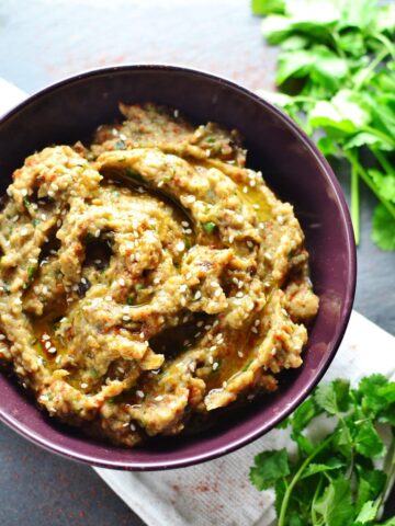 Eggplant dip in purple bowl on white cloth with fresh cilantro in top and bottom left corners.